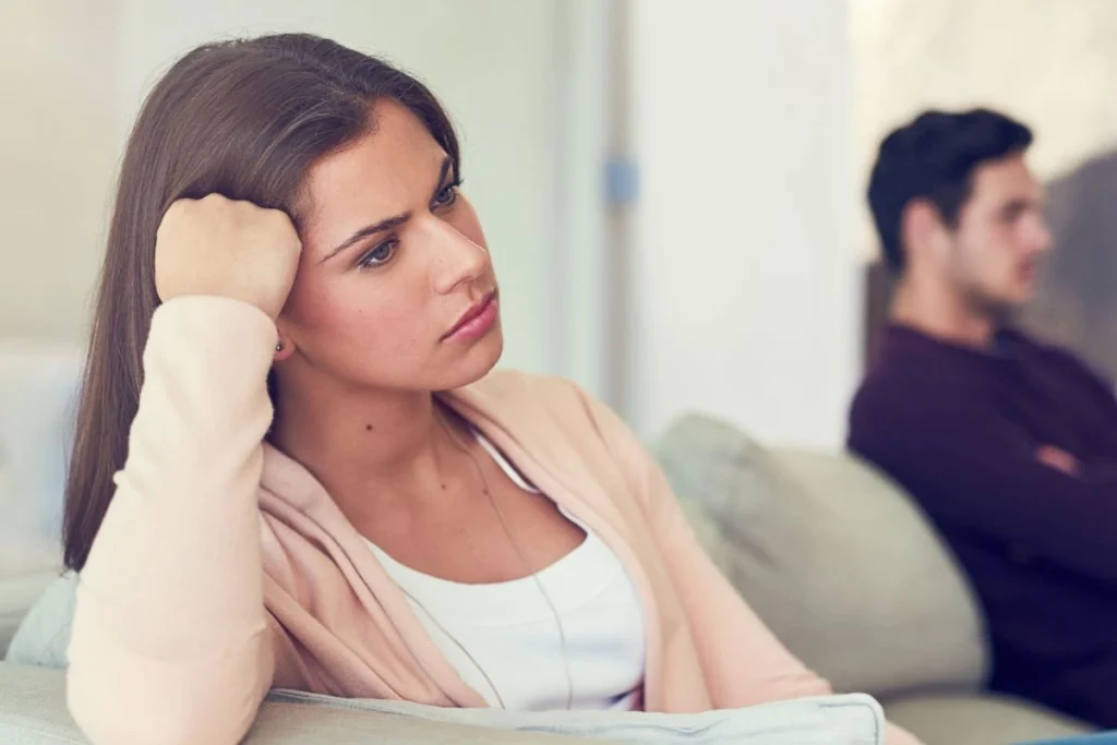 10 Early Warning Signs of Hearing Loss You Shouldn’t Ignore img A woman sits on a sofa with her head resting on her hand, looking thoughtful, as she notices the early warning signs of hearing loss; meanwhile, a man sits in the background facing away.